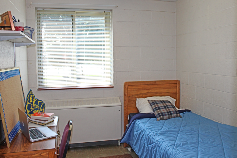 Inside of a single occupancy room inside Saffer Hall. There is a bed, desk, and shelf.